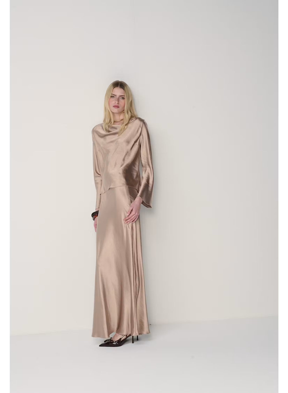 A woman with long blonde hair poses against a plain light gray background, wearing the MILVA SATIN BLOUSE IN SAND by MILVA—a shiny, champagne-beige long-sleeve blouse featuring a chic cowl neckline—paired with a matching skirt and accessorized with bracelets on her left wrist.