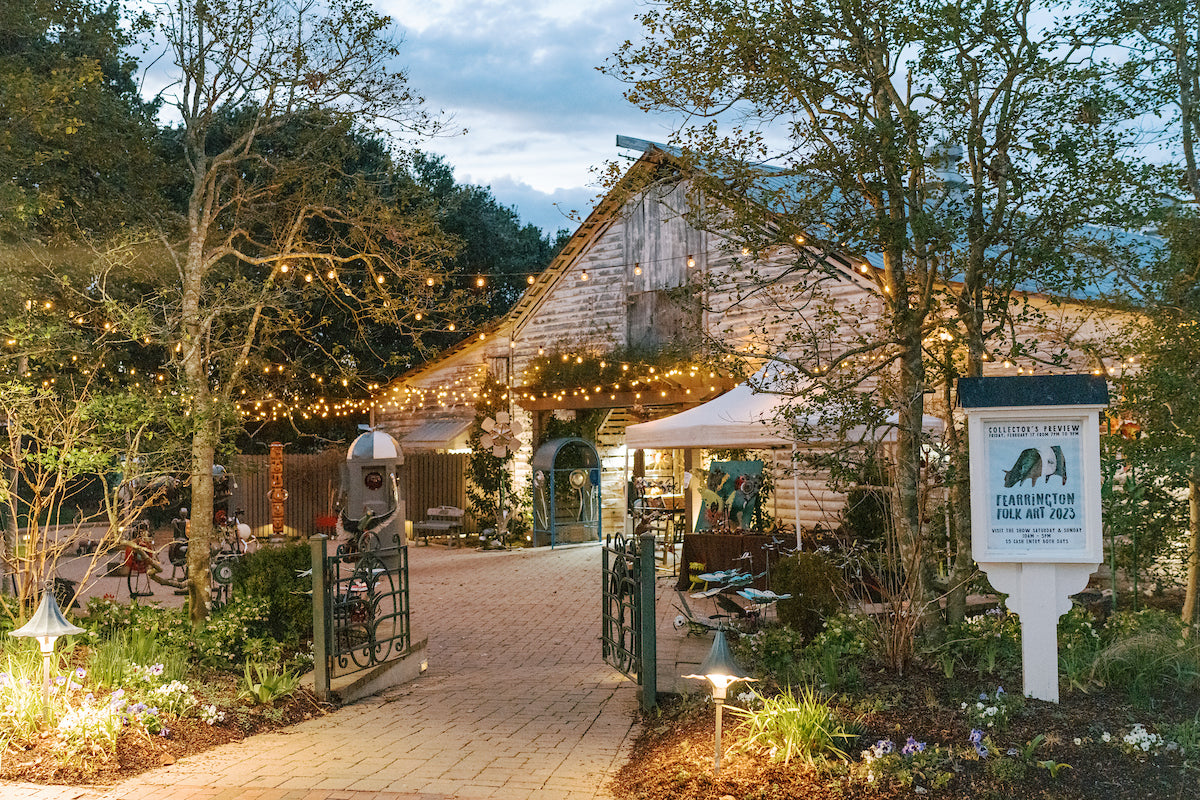 A rustic barn with string lights is surrounded by trees and gardens at dusk. The entrance gate is open, and a white sign reads “Farmington Folk Art Days.” Tables and chairs are set up outside under a tent.