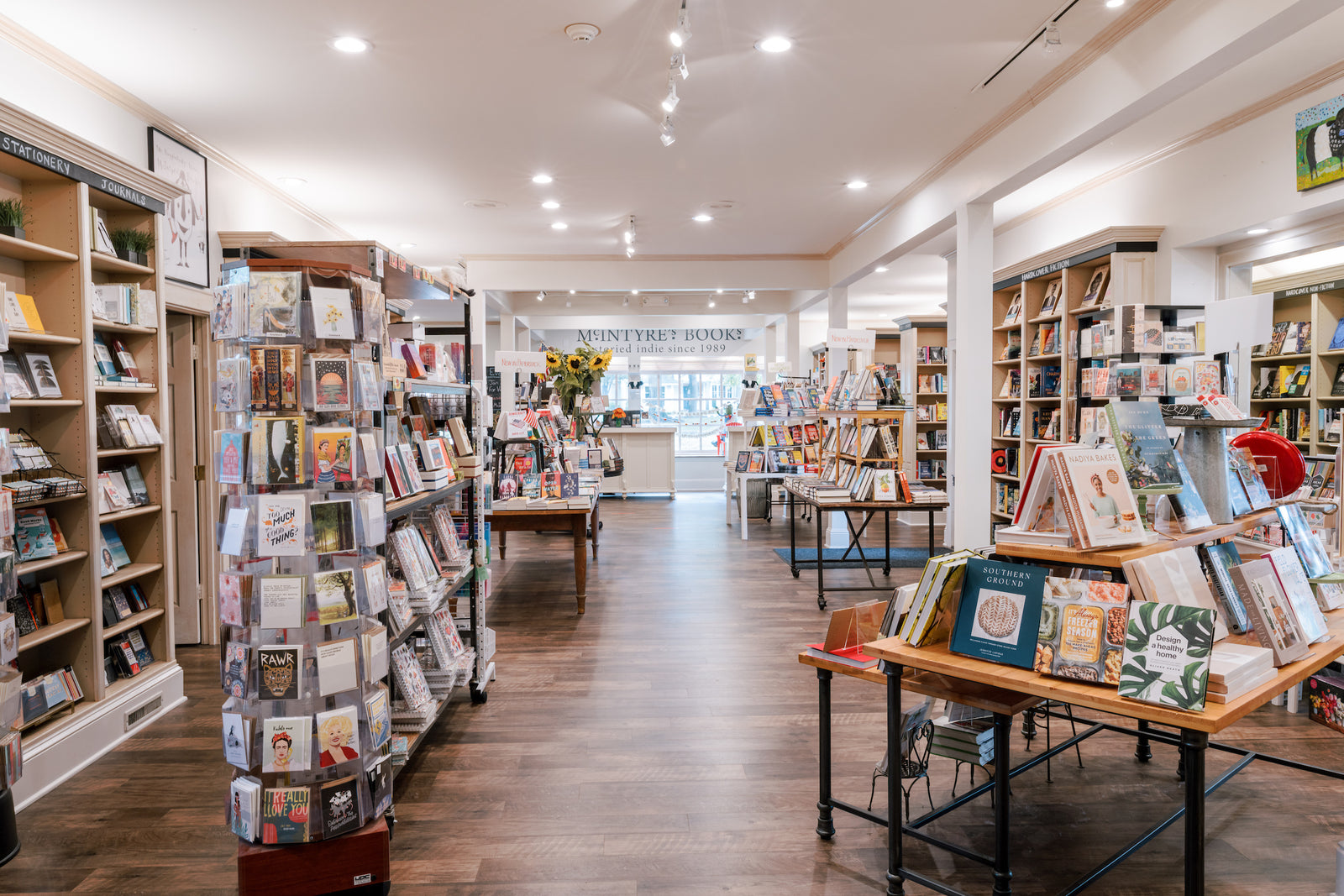 A bright, spacious bookstore with wooden floors, tall shelves filled with books, and display tables featuring assorted titles. Natural light and ceiling lights illuminate the neatly organized, inviting space.