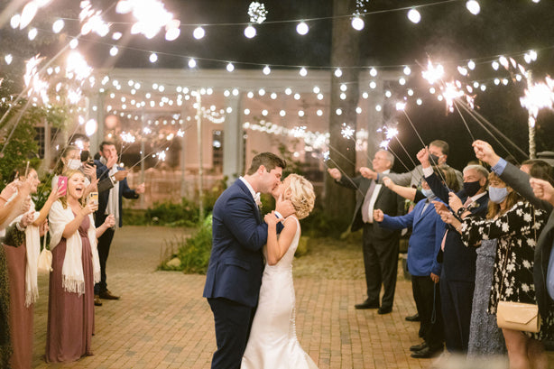 A bride and groom share a kiss under string lights as guests hold sparklers and celebrate around them at an outdoor wedding reception. Some guests wear masks, and everyone looks joyful.