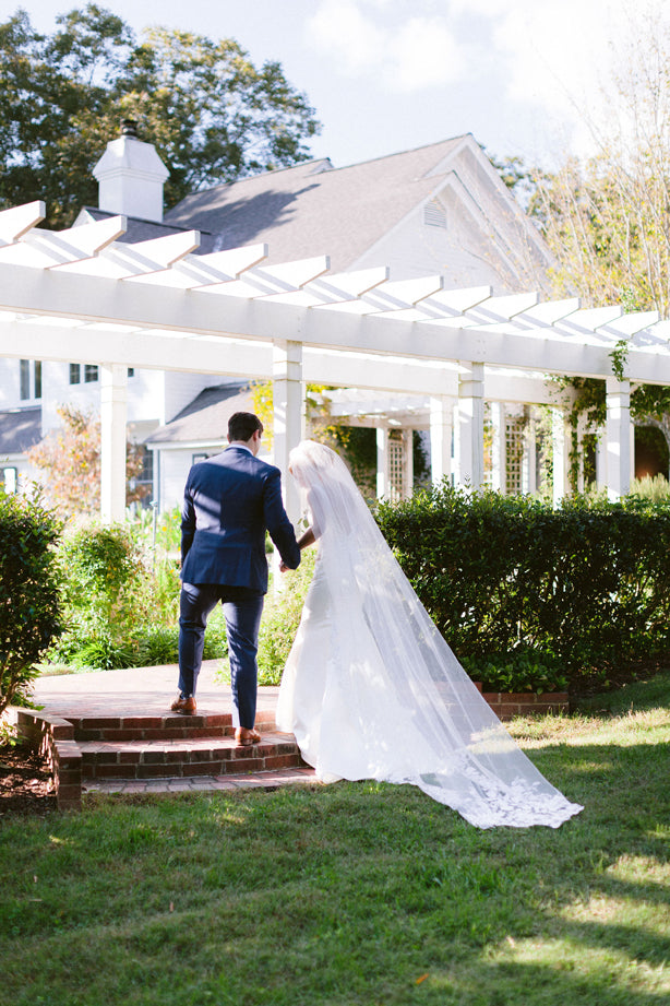 A bride in a long white gown and veil walks up brick steps beside a groom in a navy suit, heading toward a white pergola and a house in a lush garden setting.
