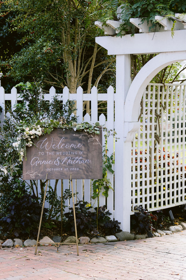 A chalkboard wedding sign with floral decorations reads “Welcome to the wedding of Annie & Nathan, October 17, 2020,” standing on an easel by a white picket fence and garden archway.