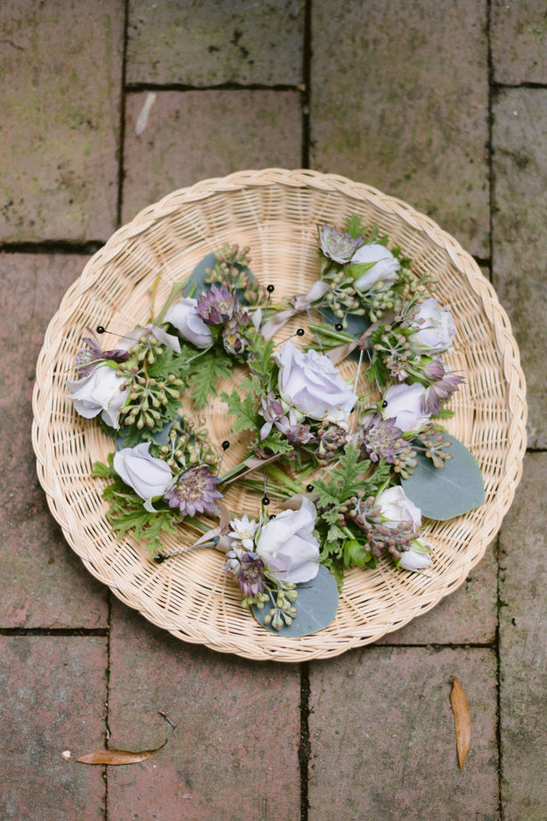 A round wicker tray containing several small floral arrangements with pale purple roses, green leaves, and purple buds, placed on a brick surface.