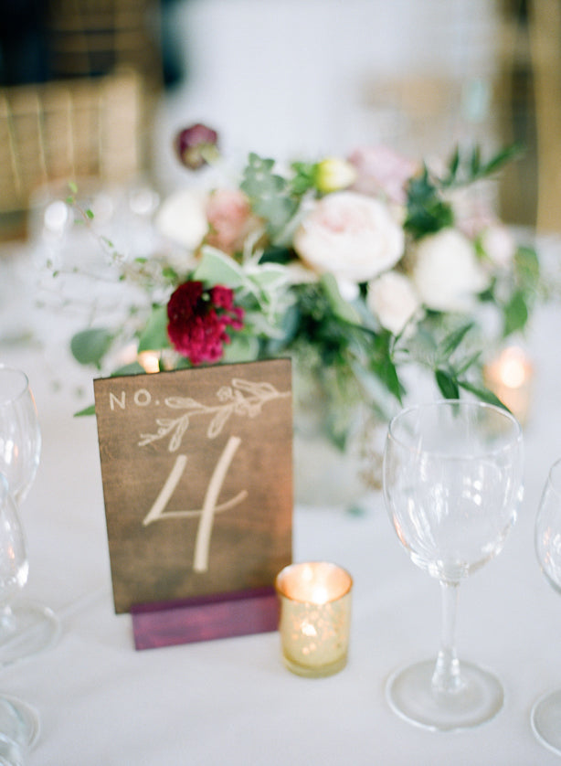 A wedding table centerpiece with a floral arrangement of white and pink flowers, a card displaying the number 4, two empty wine glasses, and lit votive candles on a white tablecloth.