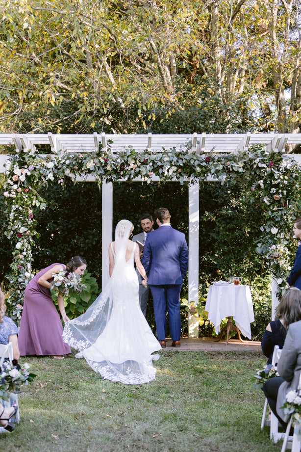 A bride in a white dress and veil walks toward a groom in a blue suit at an outdoor wedding ceremony under a floral-covered pergola, with guests seated on either side and trees in the background.