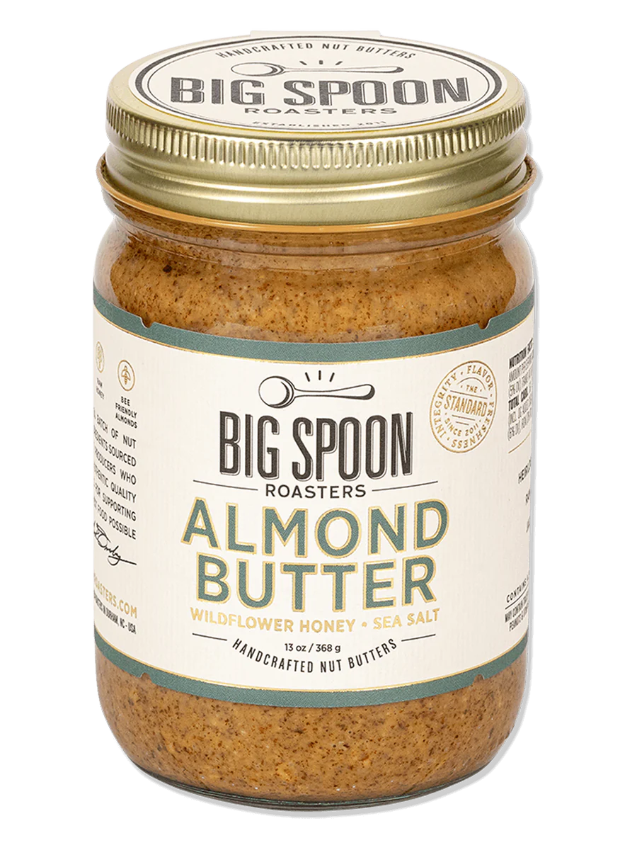 A jar of BIG SPOON ROASTERS Almond Butter with a golden lid. The label reads: &quot;Handcrafted Nut Butter,&quot; &quot;BIG SPOON ROASTERS - ALMOND BUTTER WILDFLOWER HONEY NUT BUTTER,&quot; &quot;With Organic Wildflower Honey &amp; Sea Salt,&quot; and &quot;12 oz.&quot; Featuring heirloom Mission almonds, the label boasts a spoon illustration and other decorative elements.