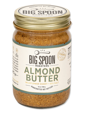 A jar of BIG SPOON ROASTERS Almond Butter with a golden lid. The label reads: "Handcrafted Nut Butter," "BIG SPOON ROASTERS - ALMOND BUTTER WILDFLOWER HONEY NUT BUTTER," "With Organic Wildflower Honey & Sea Salt," and "12 oz." Featuring heirloom Mission almonds, the label boasts a spoon illustration and other decorative elements.