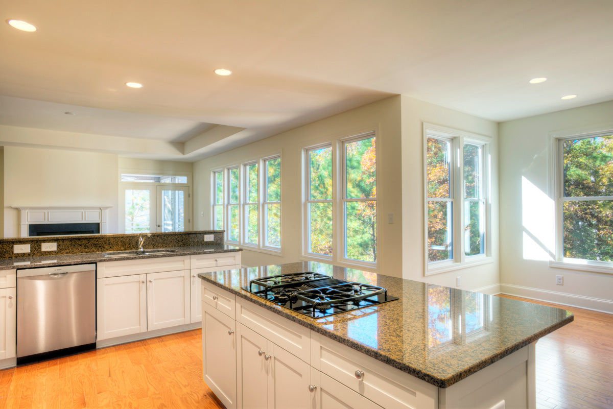 Bright modern kitchen with white cabinets, stainless steel appliances, granite countertops, a gas cooktop island, and large windows letting in abundant natural light with views of green trees outside.