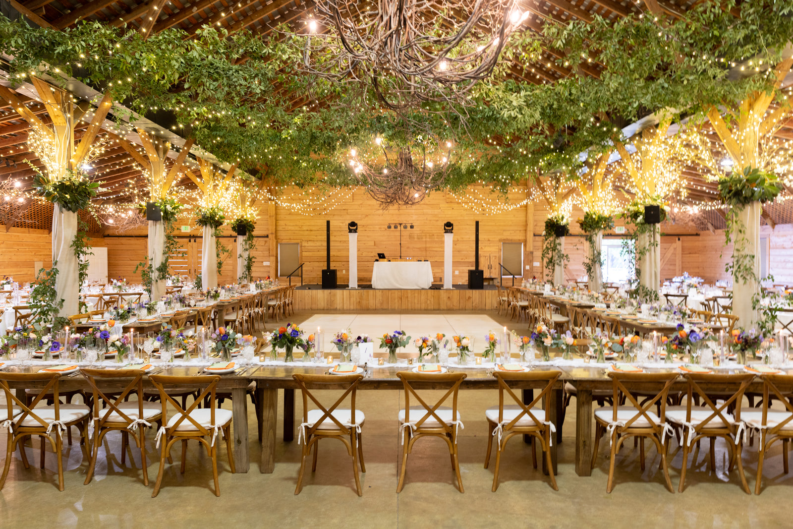 A large, rustic wedding reception hall decorated with string lights, greenery, and wooden beams. Long banquet tables with floral centerpieces surround a central dance floor, and a DJ booth stands at the far end.