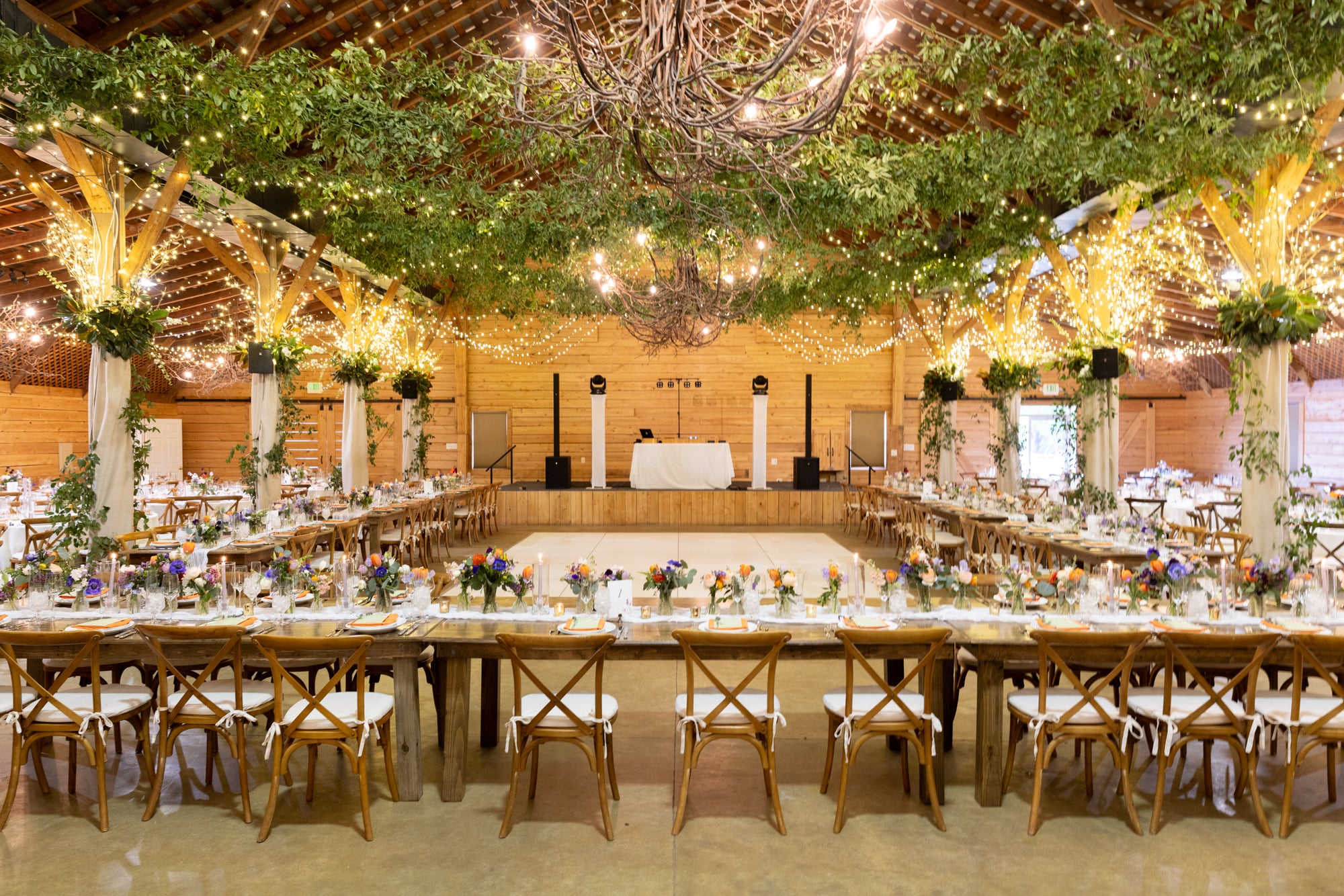 A large, rustic wedding reception hall decorated with string lights, greenery, and wooden beams. Long banquet tables with floral centerpieces surround a central dance floor, and a DJ booth stands at the far end.