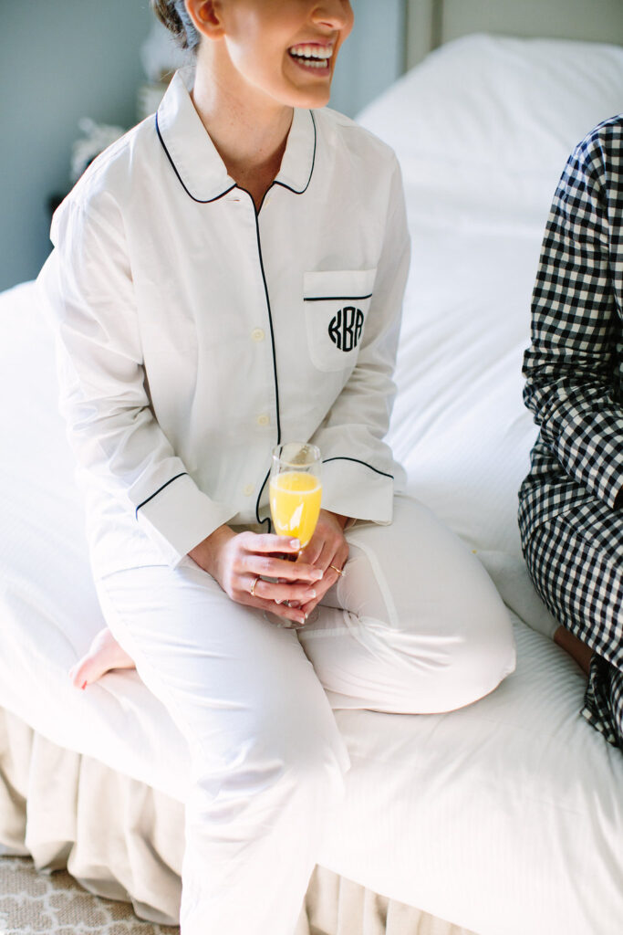 Woman in white pajamas with black trim, holding a mimosa glass and sitting on a bed, smiling. Another person in black-and-white checkered pajamas sits next to her, partially out of frame.