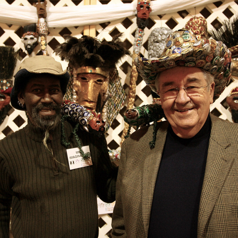 Two smiling men pose together at an art display. One wears a dark hat and sweater, the other a jacket and a hat decorated with colorful bottle caps. Ornate masks are displayed in the background.