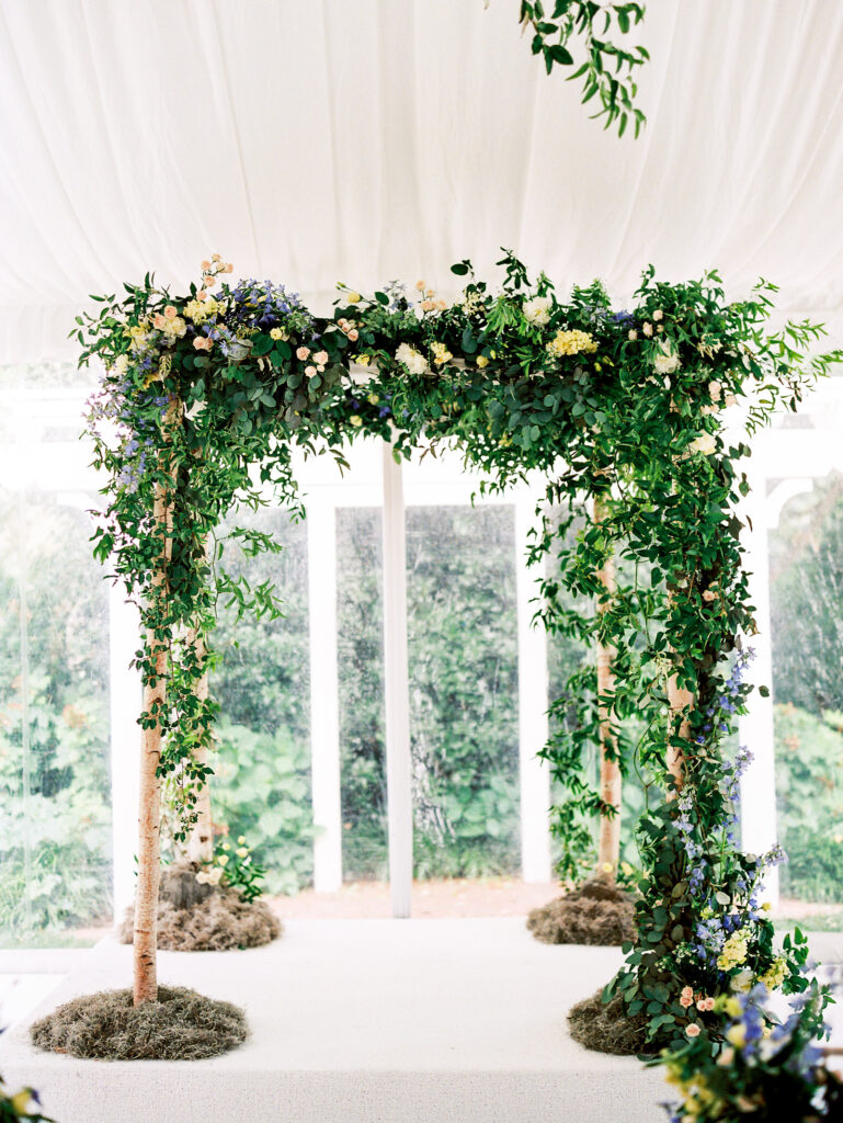 A wedding arch made of birch branches and lush greenery with white and purple flowers stands under a white tent with natural light and leafy plants in the background.