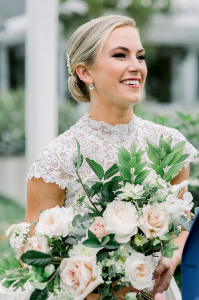 A smiling bride in a lace wedding dress holds a large bouquet of white and blush flowers with greenery. She wears pearl earrings and her hair is styled in an elegant updo.