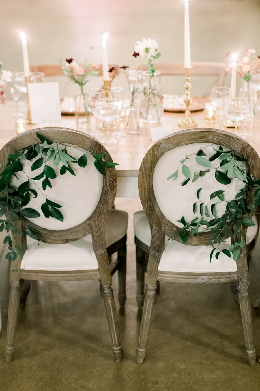Two wooden chairs with white cushions are decorated with green leafy branches on their backs, facing a table set with candles, flowers, and glassware, creating an elegant, romantic atmosphere.
