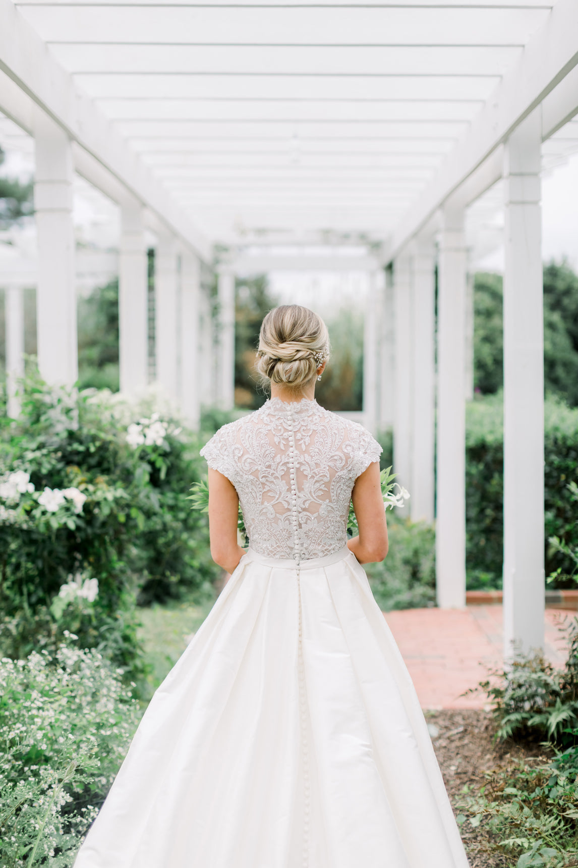 A bride stands outdoors with her back to the camera, wearing a white gown with lace detailing and cap sleeves. She is under a white pergola surrounded by greenery and white flowers. Her hair is styled in an elegant updo.