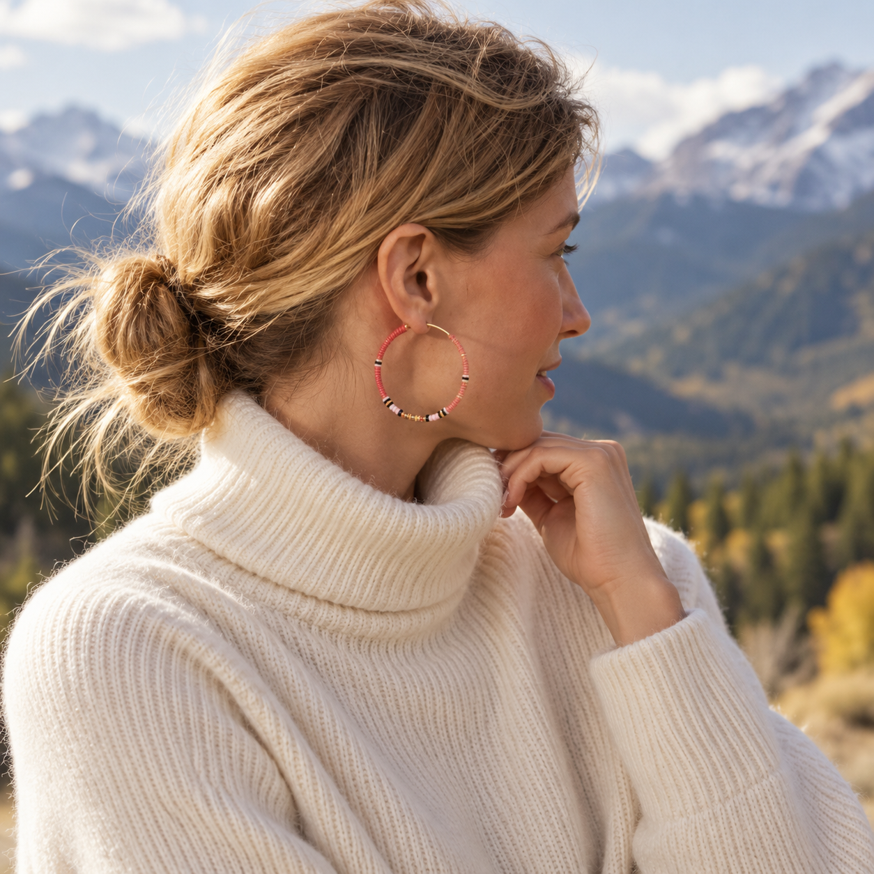 A woman in a cream turtleneck sweater wearing a gold hoop earring with pink and black beads, with a mountain landscape in the background.