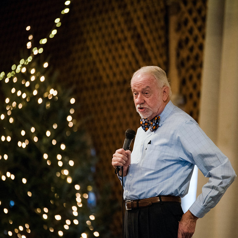 An older man with a white beard, wearing a blue shirt and a colorful bow tie, holds a microphone while speaking. A decorated, lit Christmas tree is visible in the background.