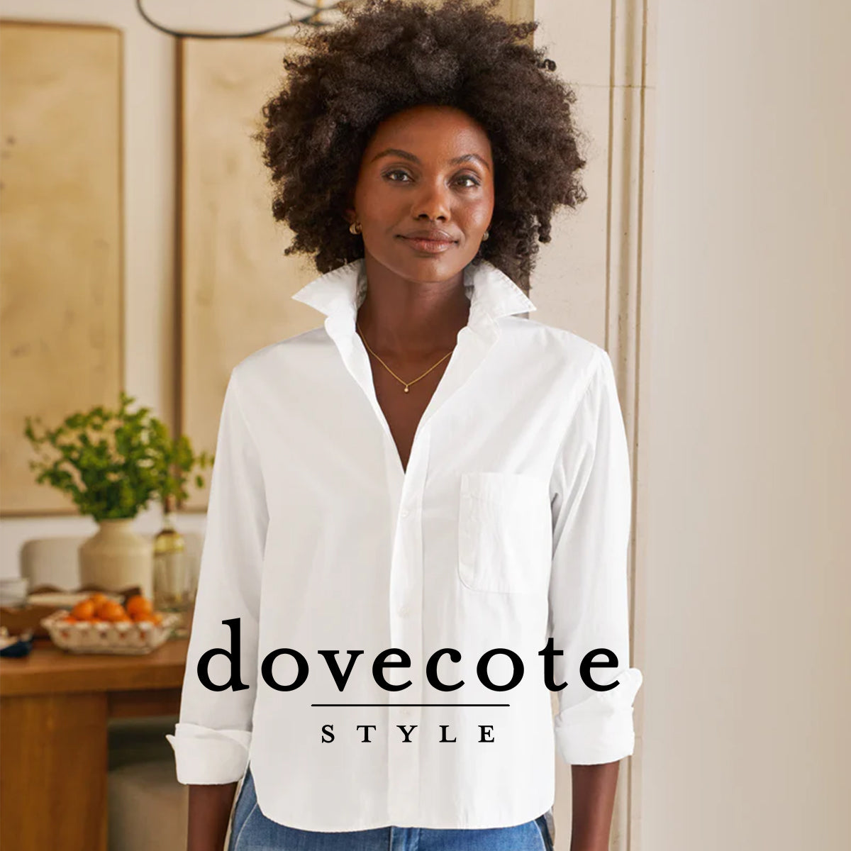 A woman with natural curly hair wearing a white collared shirt and blue jeans stands indoors, smiling gently. Text reads "dovecote STYLE." A dining table with fruit and a green plant are in the background.