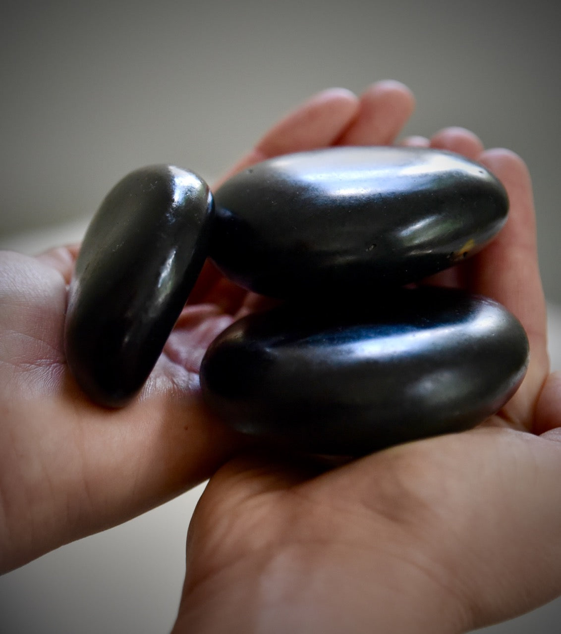 A close-up of two hands gently holding three smooth, oval-shaped black stones, with a soft, blurred background.