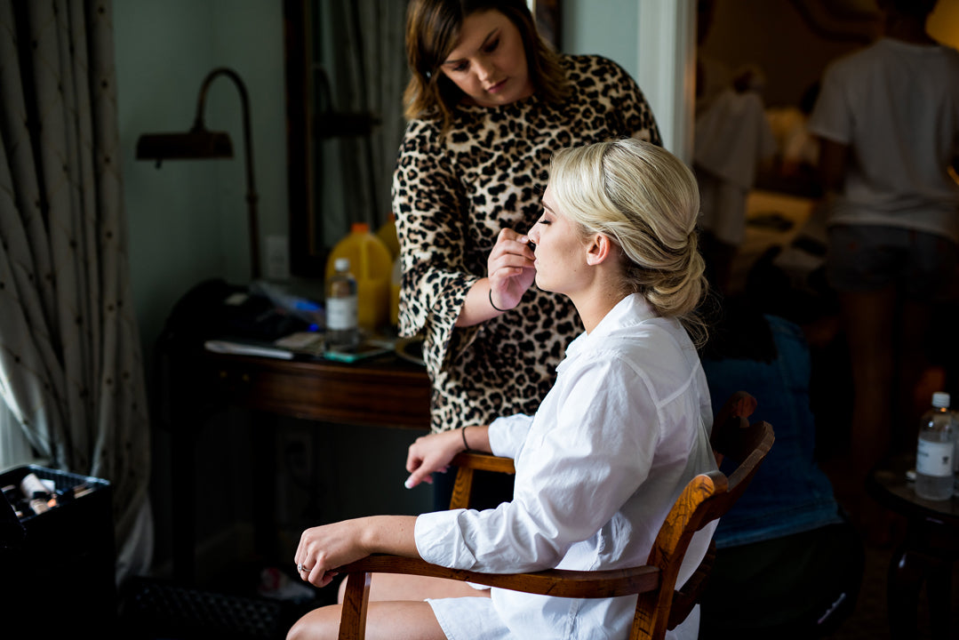 A woman in a leopard print dress applies makeup to a seated blonde woman wearing a white robe in a well-lit room, with a desk and people in the background.
