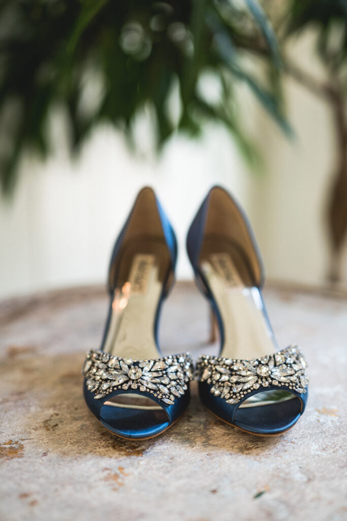 A pair of elegant blue open-toe heels with sparkling rhinestone embellishments on the front, displayed on a stone surface with blurred greenery in the background.