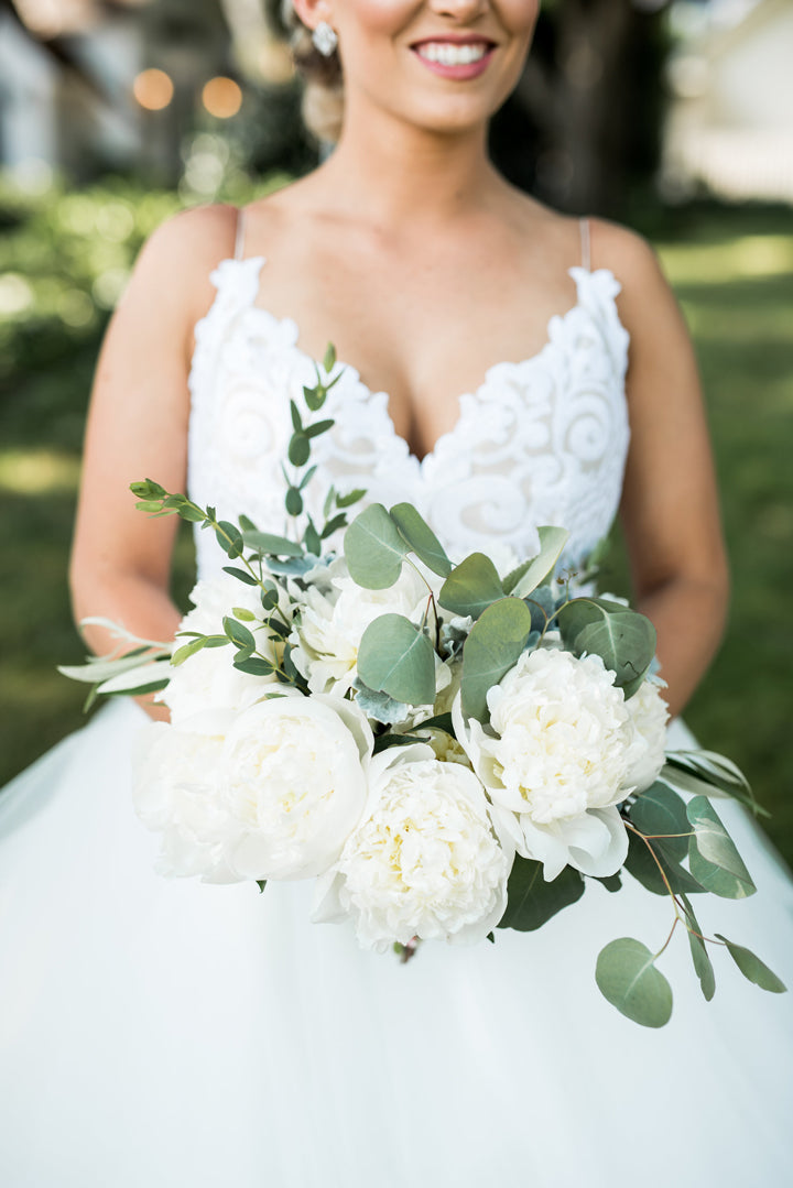 A bride in a white dress holds a bouquet of white peonies and greenery, smiling outdoors in a sunlit garden.