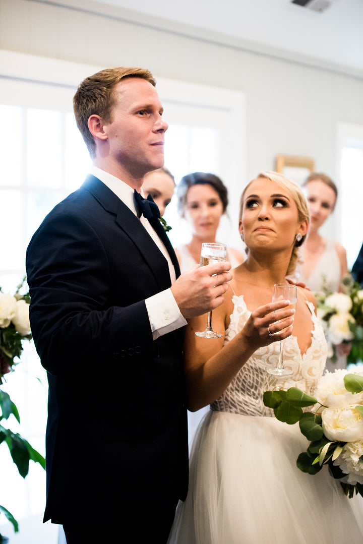 A groom in a black tuxedo and a bride in a white dress hold champagne glasses, appearing emotional during a wedding toast, surrounded by bridesmaids with bouquets in a bright indoor setting.