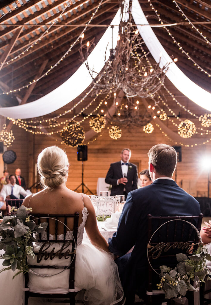 A bride and groom sit facing a speaker at their wedding reception in a rustic barn decorated with string lights, draped fabric, and elegant greenery on their chairs.