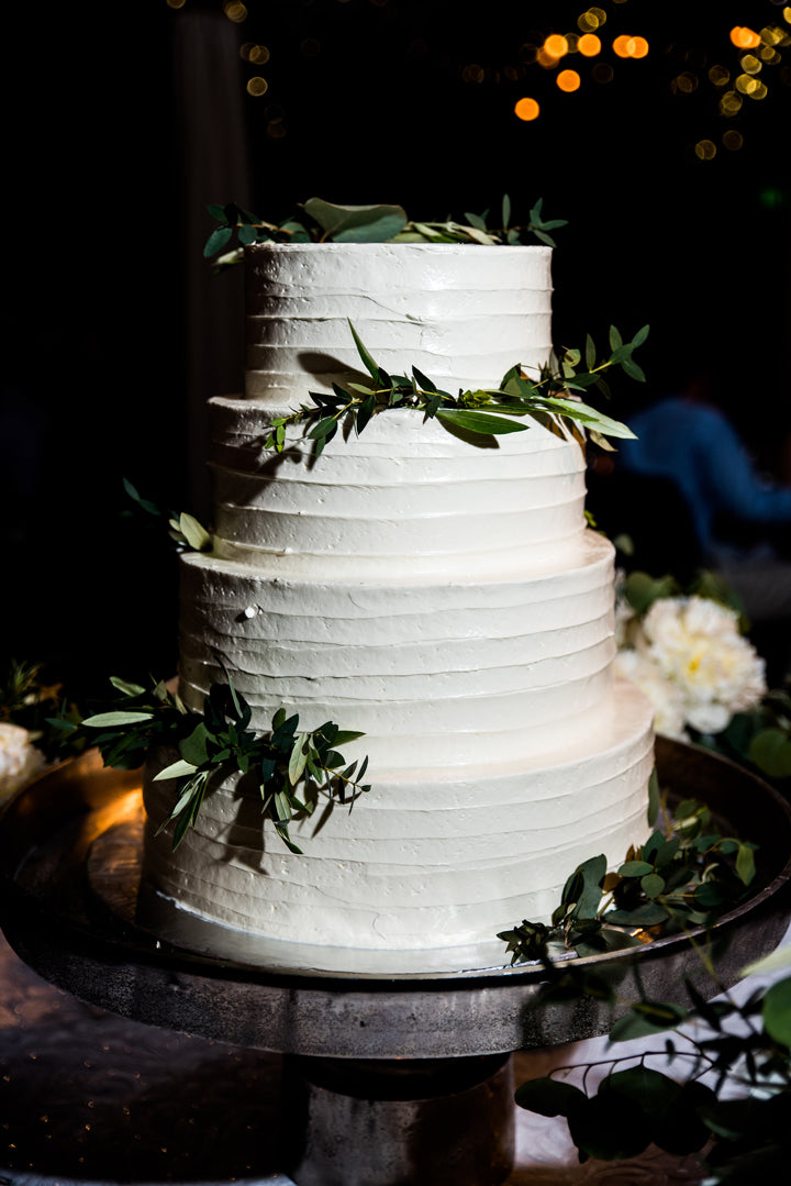 A three-tiered white wedding cake decorated with green leaves sits on a silver stand. Blurred lights and flower arrangements are visible in the dark background.