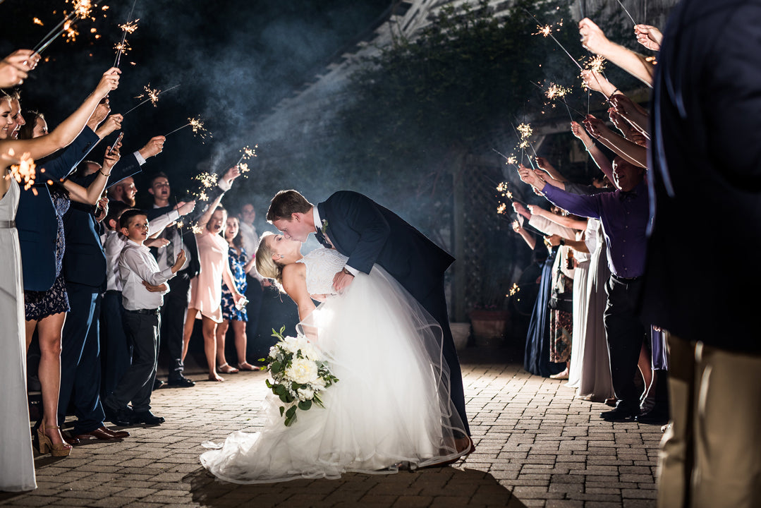 A bride and groom share a kiss as the groom dips the bride; guests on both sides hold sparklers overhead, lining a walkway at night and celebrating the couple’s wedding.