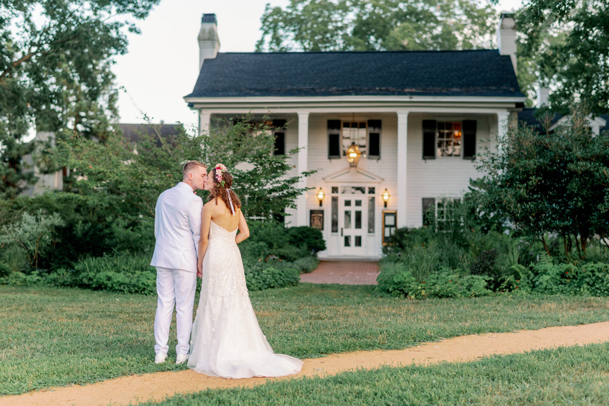 A bride and groom stand close together, facing a large white house with black shutters and glowing lights, surrounded by lush greenery and gardens. The bride wears a white gown; the groom wears a white suit.