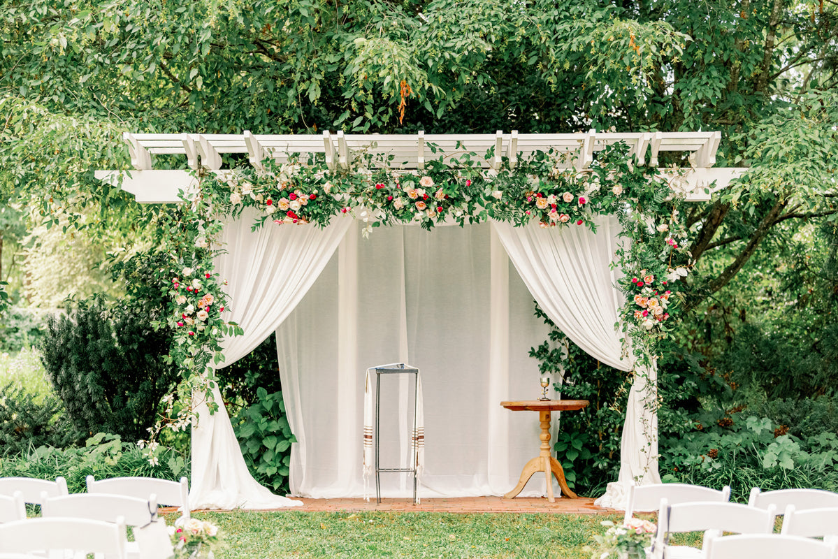A white wedding arch decorated with lush greenery and pink flowers, draped with white curtains, stands outdoors on grass. A small table and a metallic stand are beneath the arch, with trees in the background.