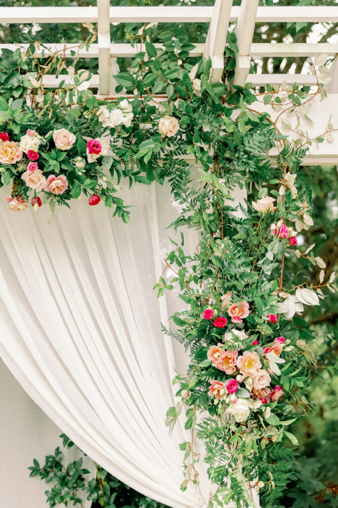 A floral wedding arch decorated with lush greenery, white drapery, and clusters of pink, red, and cream roses, set outdoors under a white pergola.