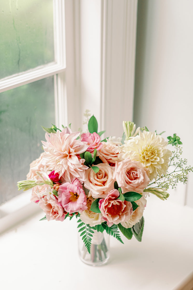 A bouquet of pink and cream roses, dahlias, and greenery in a glass vase sits on a white surface near a window with condensation.