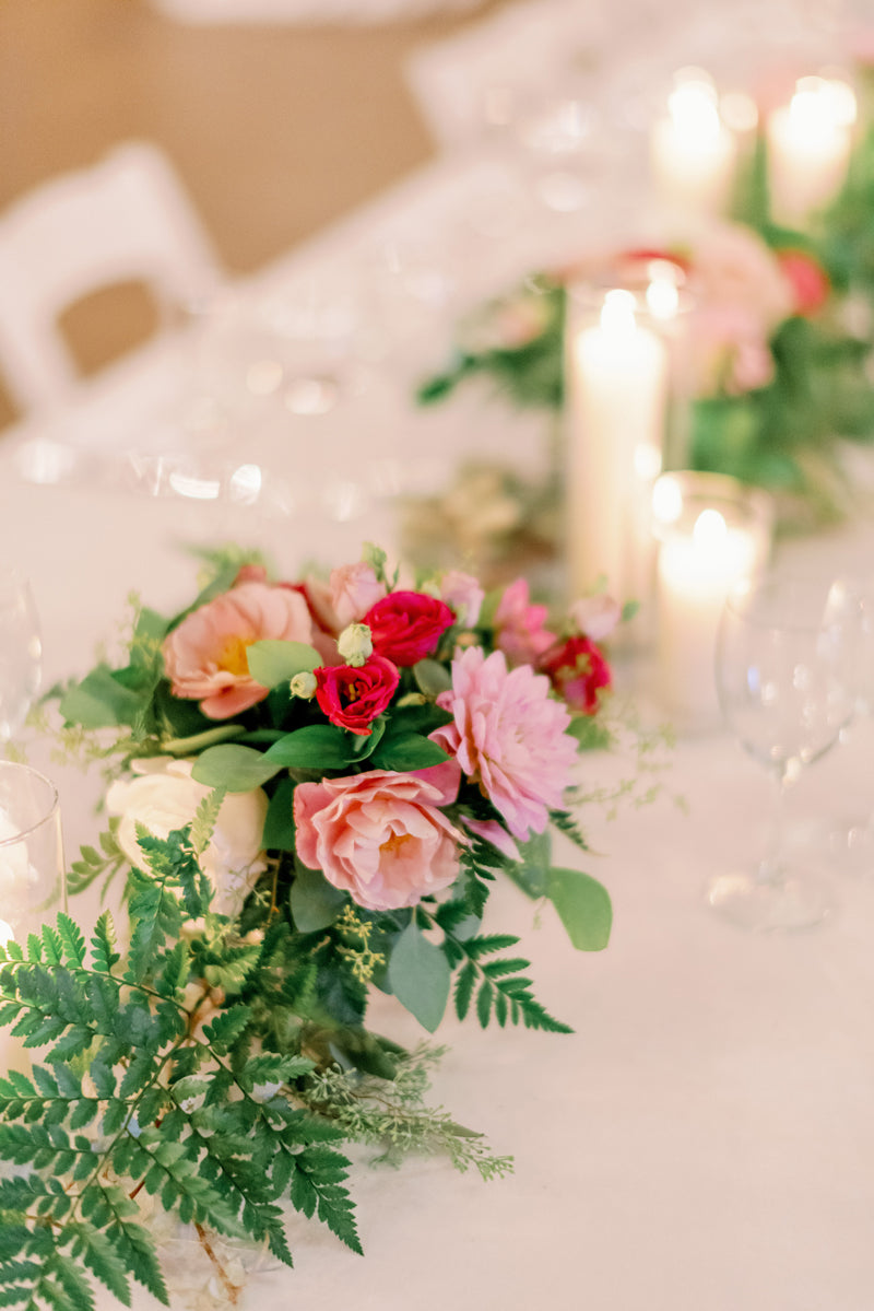 A close-up of an elegant table centerpiece featuring pink and red flowers, green ferns, and greenery, surrounded by glowing white candles and wine glasses on a white tablecloth.