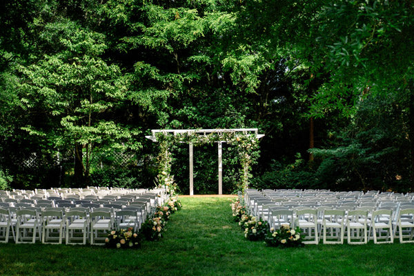 An outdoor wedding ceremony setup with rows of white chairs facing a flower-decorated arbor, surrounded by lush green trees and greenery, on a sunny day.
