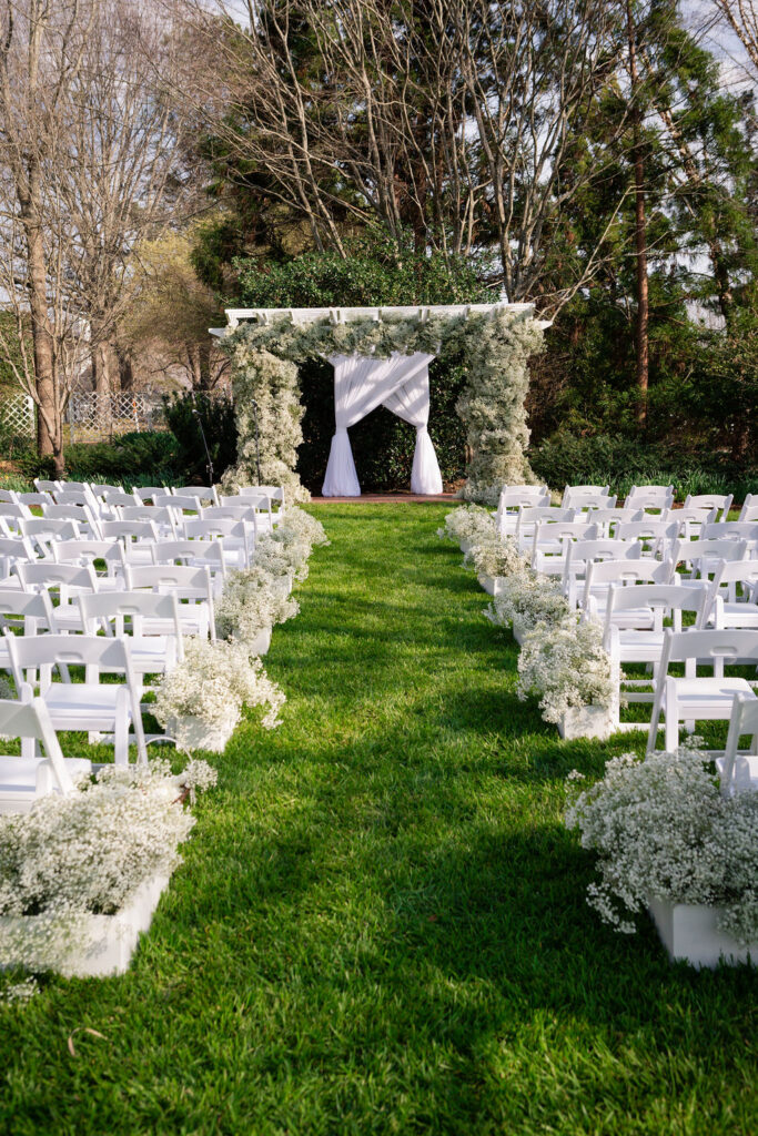 A grassy outdoor aisle lined with white chairs and floral arrangements leads to a white wedding arch decorated with flowers and draped fabric, set amidst trees and greenery.