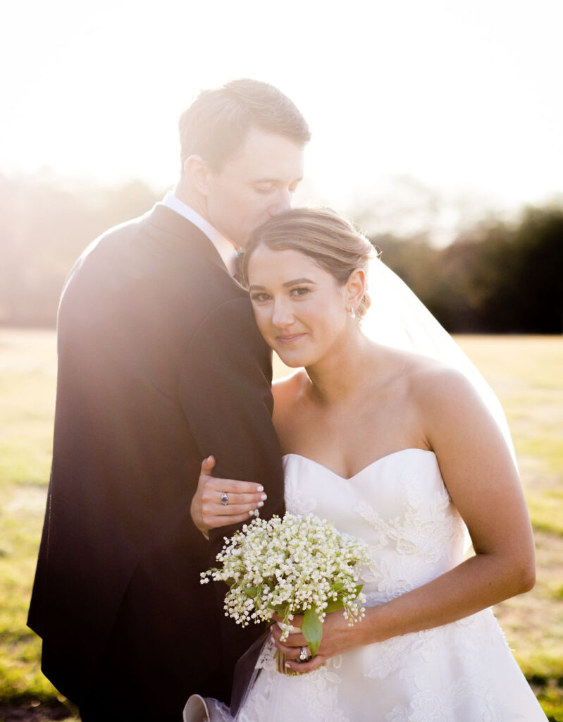 A groom in a black suit gently kisses the bride’s forehead as she smiles and holds a small bouquet of white flowers outdoors, bathed in warm sunlight.