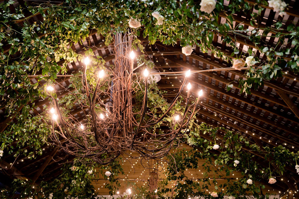A rustic chandelier with glowing bulbs hangs from a wooden ceiling, surrounded by green foliage, vines, and white roses. Warm string lights add a cozy, festive ambiance to the scene.