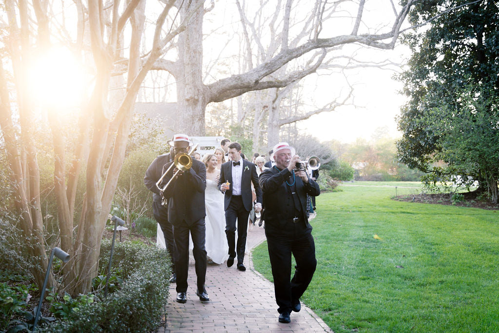 A wedding procession follows a small band playing brass instruments along a brick path in a garden, with sunlight streaming through bare trees.