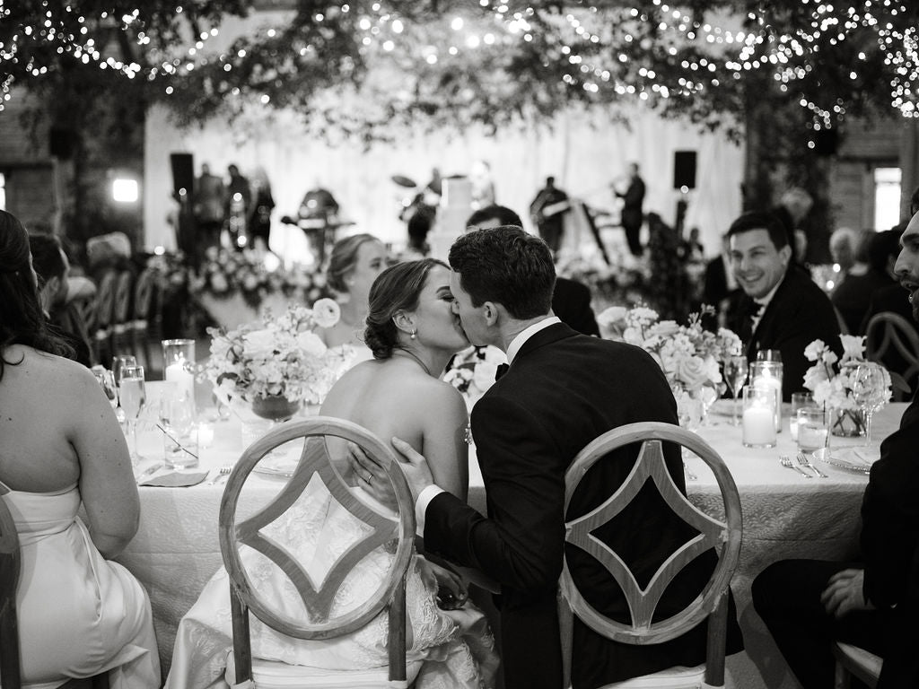A bride and groom share a kiss while seated at a decorated table during their wedding reception. Guests sit around them, and twinkling lights and a live band are visible in the background.