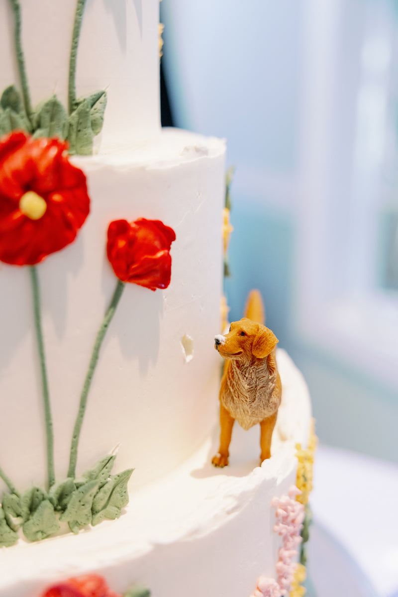 A close-up of a white, tiered cake decorated with red and pink flowers, featuring a small, realistic dog figurine standing beside one of the tiers.