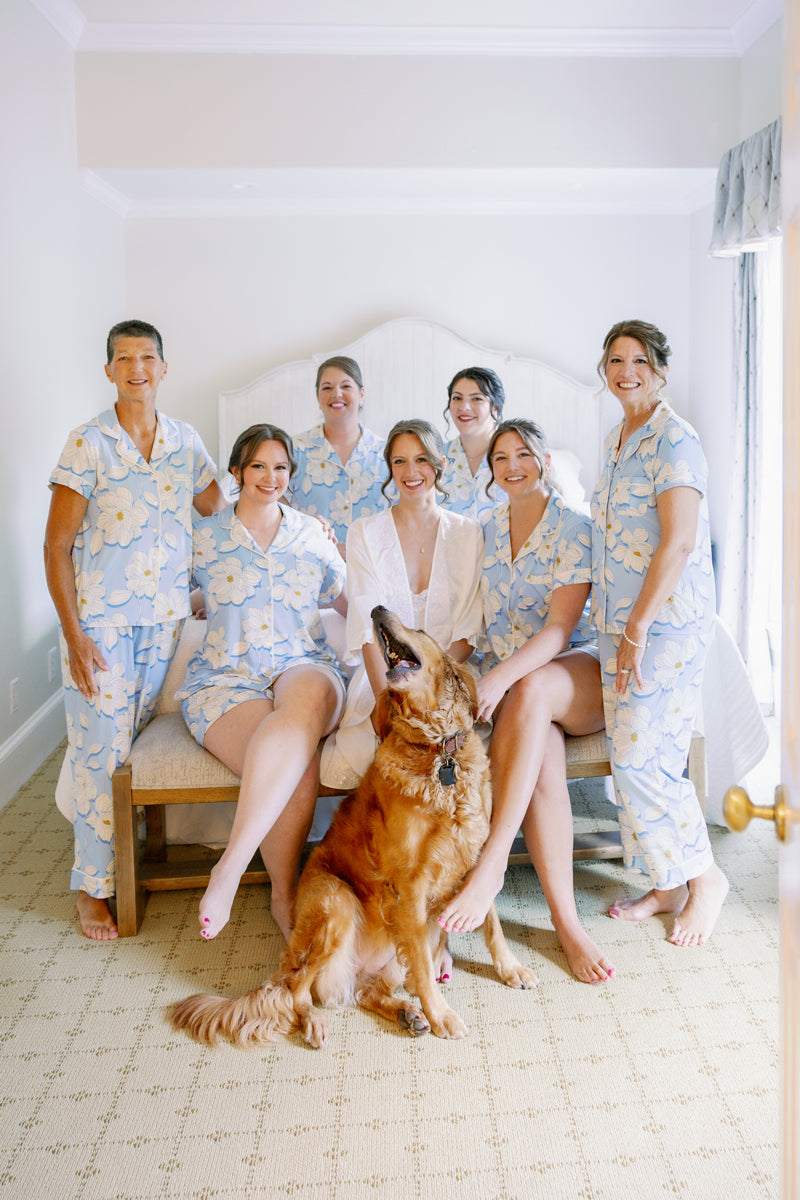 A bride in white sits on a bed surrounded by five women in matching blue floral pajamas and a golden retriever sitting on the floor in front of them, all smiling in a bright, cozy room.