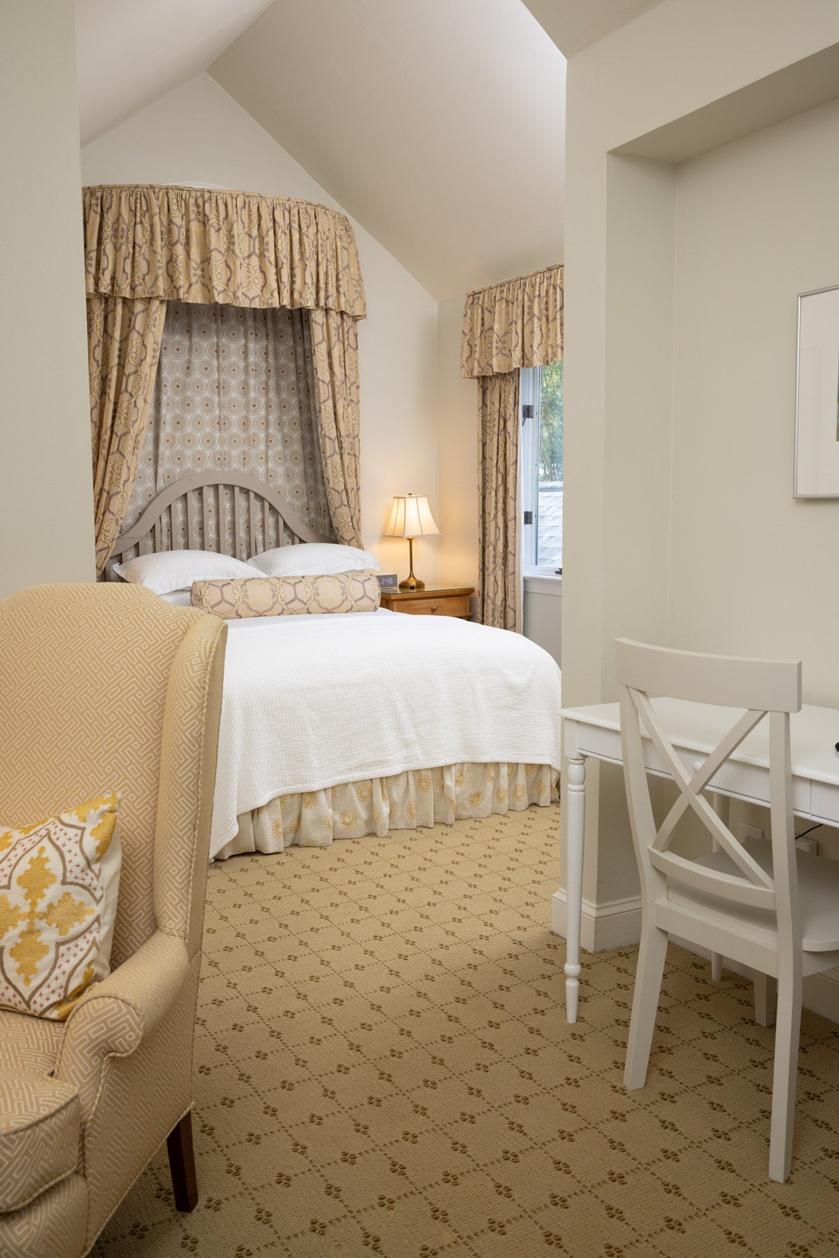 A cozy bedroom with a white bed, matching patterned curtains, a small bedside lamp, a white desk with a chair, and a beige armchair with a yellow cushion on a carpeted floor.