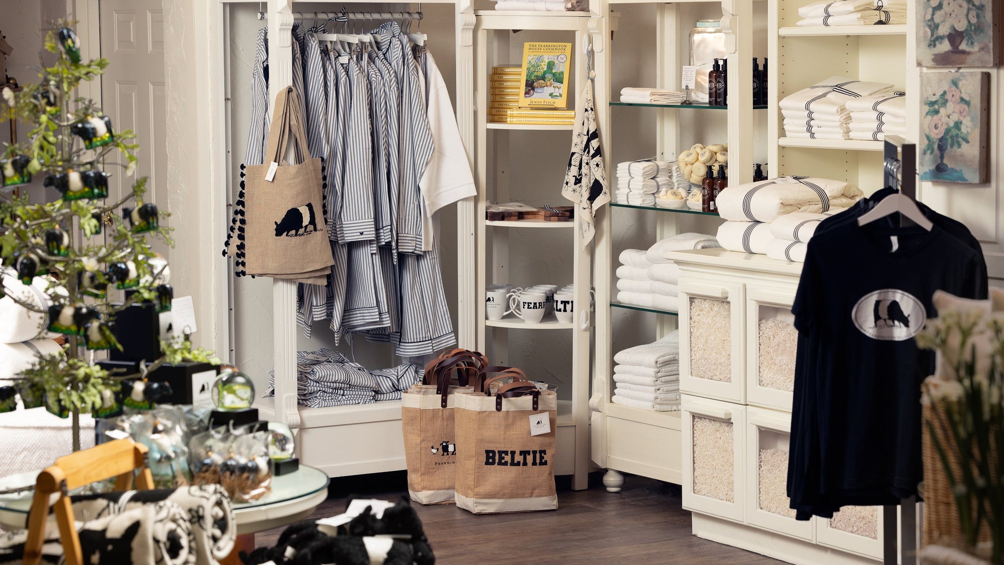 A cozy clothing and gift shop displays pajamas, towels, shirts, tote bags, and home decor on white shelves and racks. A small decorated Christmas tree and various gift items are visible on tables.