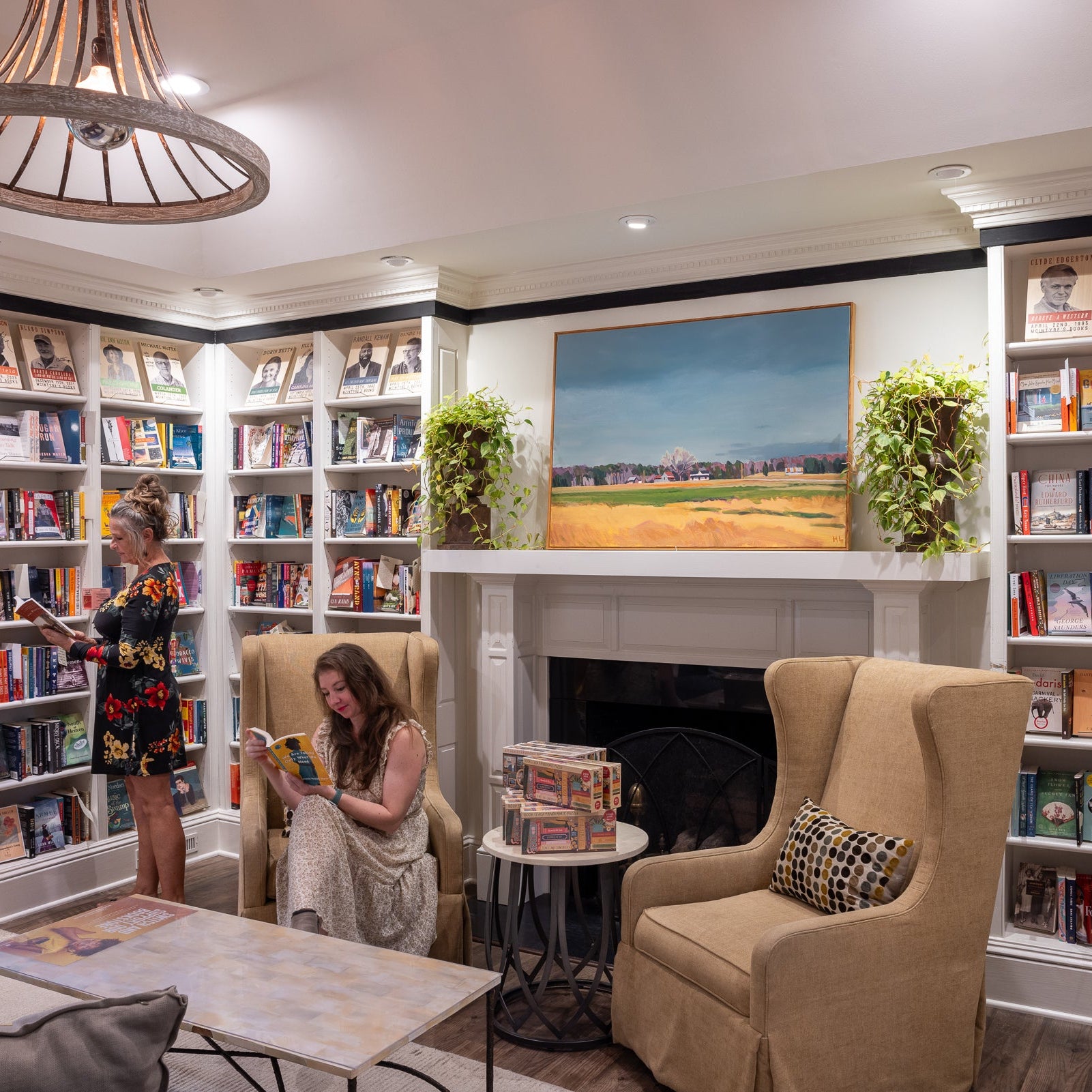 Two women in a cozy library: one stands browsing a bookshelf, the other sits reading in a beige armchair. The room has bookshelves, a fireplace with plants, a landscape painting, and warm lighting.