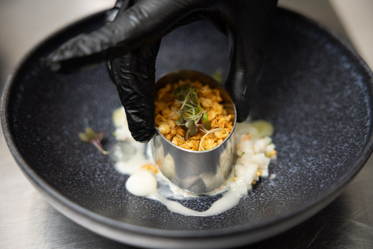A gloved hand places a metal ring mold filled with a crumbled food mixture onto a dark plate, garnished with herbs and surrounded by white sauce and small vegetable pieces.