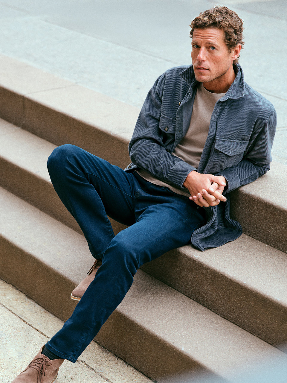 A man sits on outdoor steps wearing a gray corduroy jacket over a beige shirt, navy pants, and brown shoes. He has short curly hair and is looking slightly to the side with his hands clasped.
