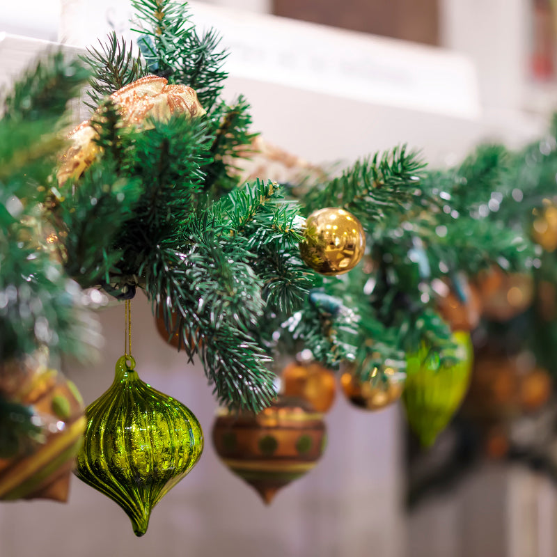 Close-up of a festive green garland decorated with shiny gold and green ornaments, including a large green ornament in the foreground, hanging indoors with a blurred background.